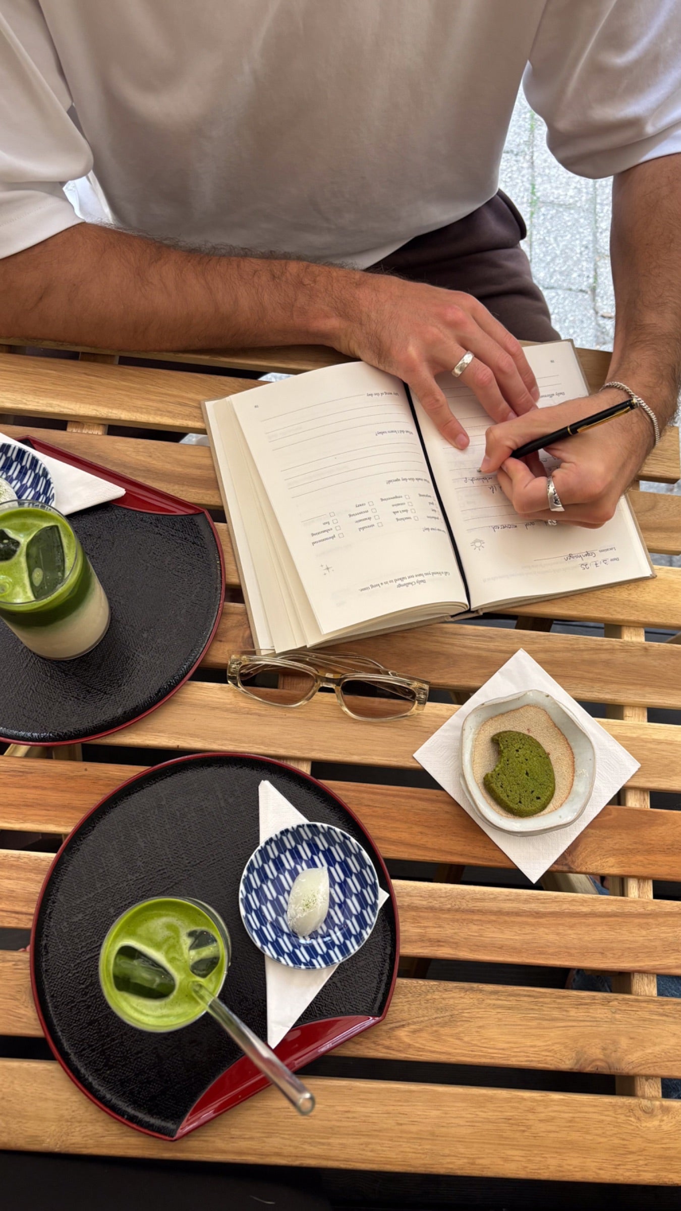 A man writes in his journal at a wooden cafe table, an iced matcha on a tray beside him