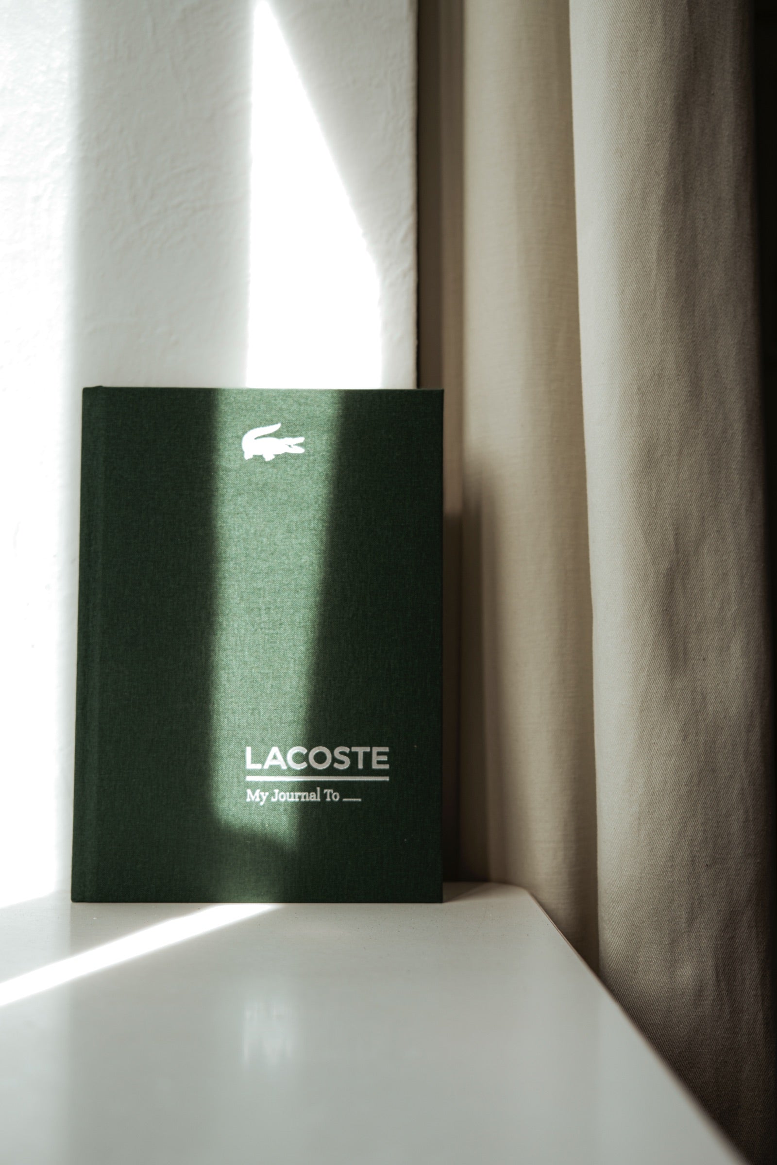 A deep green linen journal with the Lacoste crest, standing upright against a curtain in a beam of soft daylight