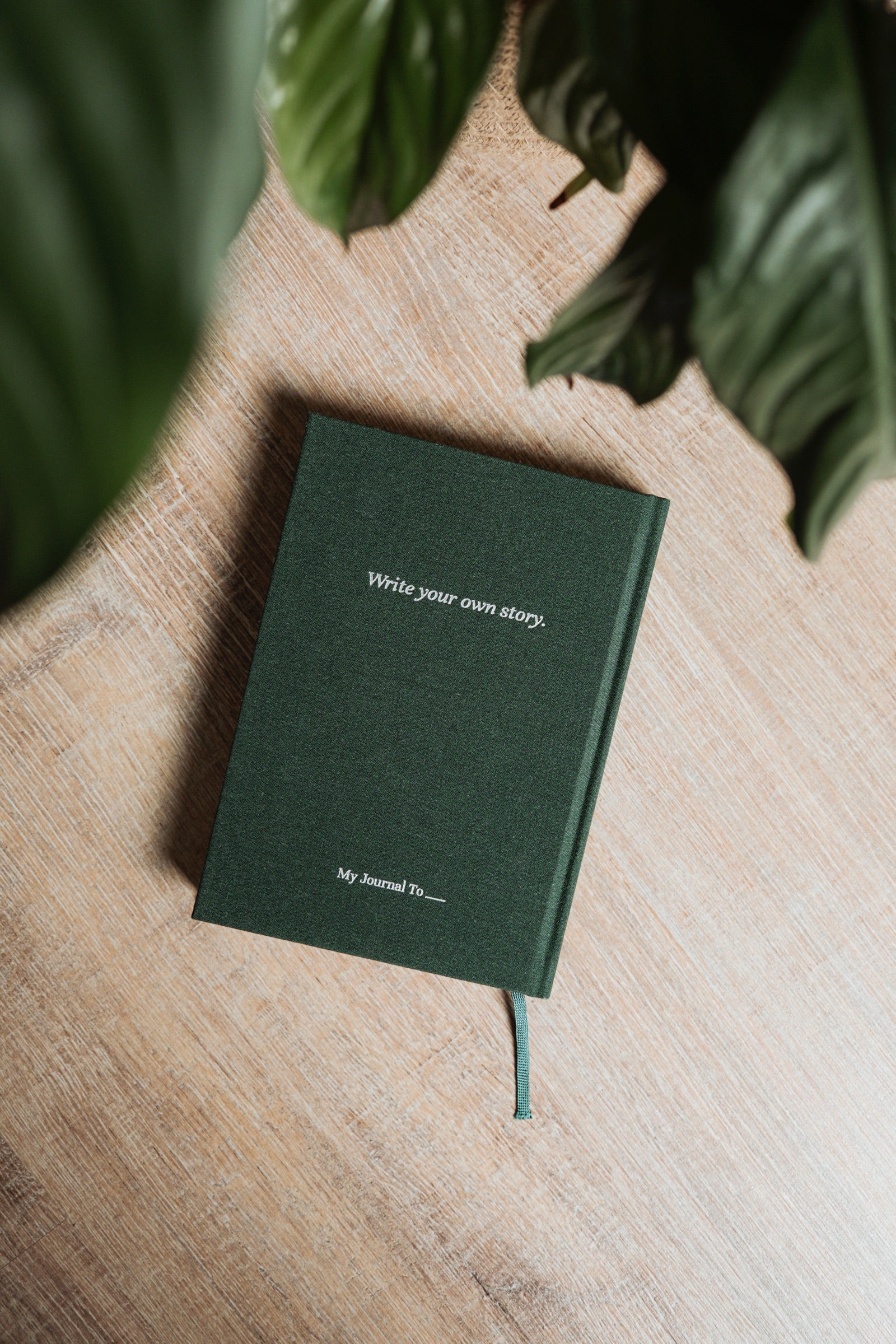 A green linen journal with the embossed line Write your own story, seen from above on a wooden surface beside plant leaves